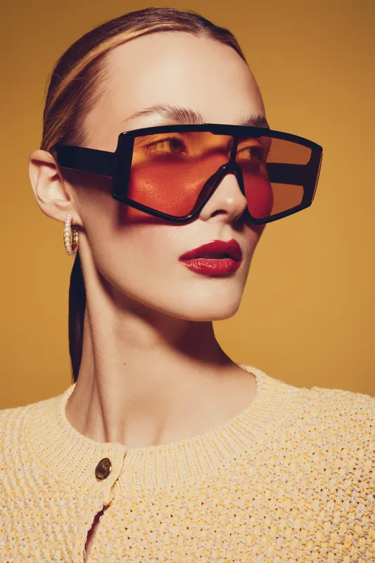Stylish woman with slicked back hair wears oversized orange sunglasses and red lipstick. She poses against a warm yellow background wearing a textured beige sweater. Fashion and beauty concept.