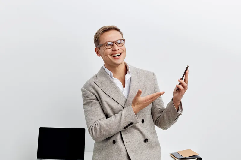 Smiling happy middle-aged man wearing glasses and a gray blazer holding a smartphone with a friendly expression, sitting at a simple workspace with a black chair and notebook, white background studio 