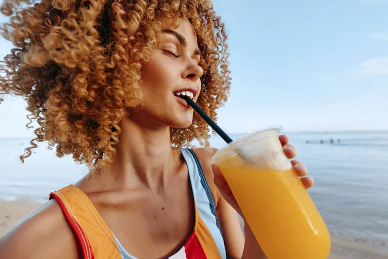 Curly haired woman on the beach sipping orange smoothie through a straw, smiling eyes closed, summer hydration moment, light wind, calm seaside background, healthy lifestyle concept
