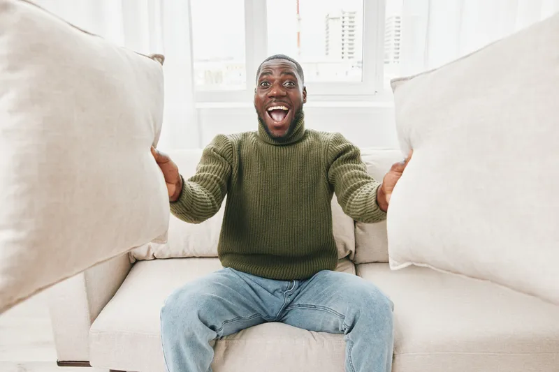 Happy man in a cozy sweater holding pillows in a bright living room, showcasing joy and comfort during a relaxed indoor moment