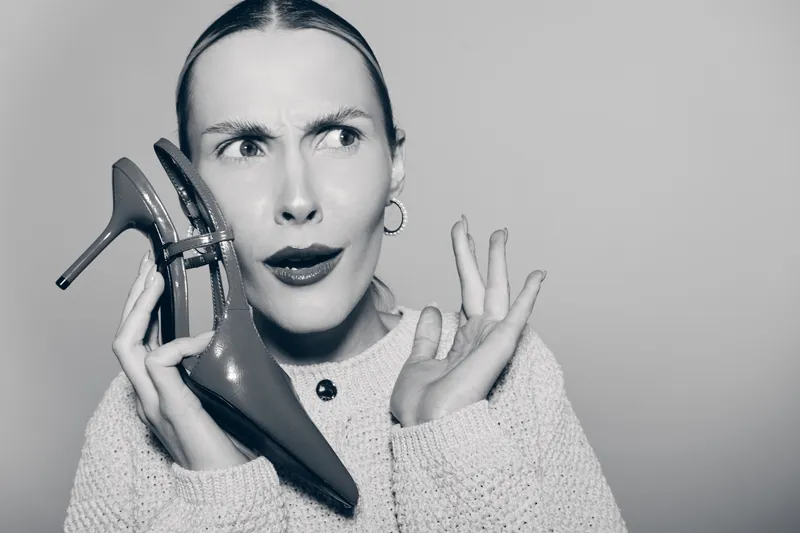Confused Caucasian woman with pale skin holds high heel shoe near face, wearing knit sweater and hoop earrings. Concept for fashion, doubt, and style choices.