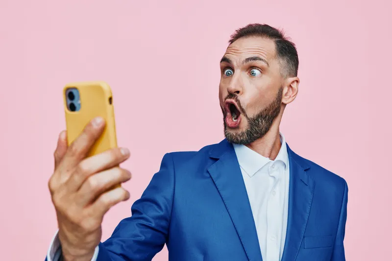 Professional man in a blue suit on a pink background, isolated setting, expressing a strong emotion. He takes a selfie with wide eyes and an open mouth, showcasing surprise and confidence.