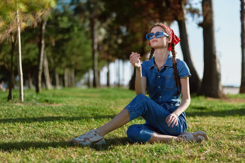 Lifestyle woman in modern boho-western denim street style sitting on grass enjoying sunny day, wearing blue sunglasses and red bandana, film color effect.