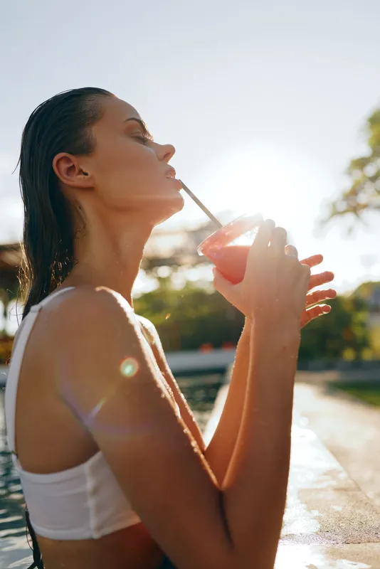 Young woman with wet hair wearing white top drinking smoothie outdoors by the pool during sunny day. Concept of healthy lifestyle, relaxation, and summer wellness.