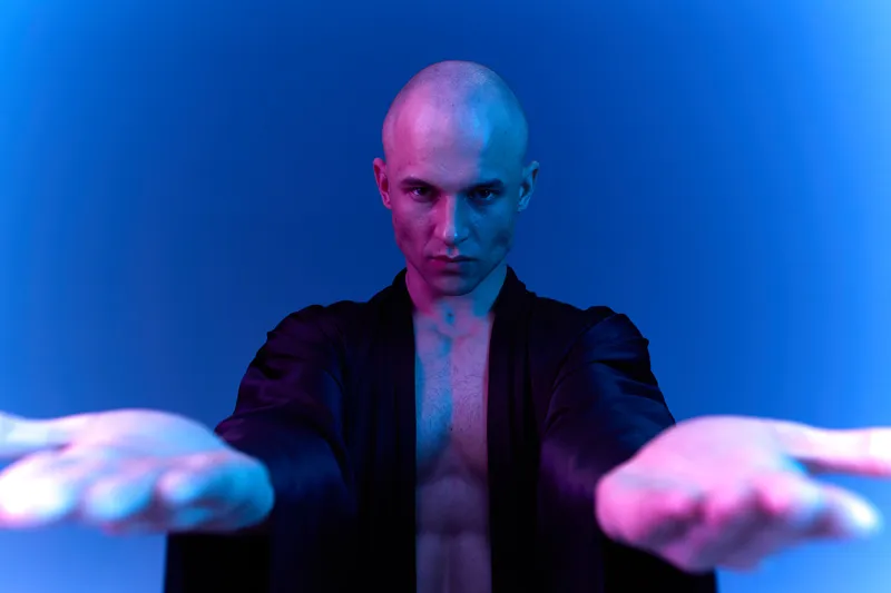 A striking portrait of a man with a bald head, extending his hands towards the viewer, illuminated by vibrant blue and purple lighting, creating a captivating atmosphere.