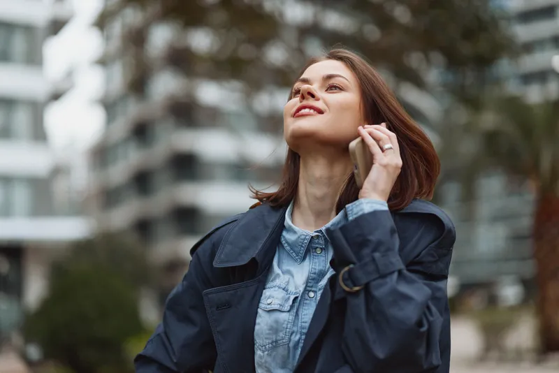 Bright woman outdoors, talking on phone, smiling face, denim shirt under a dark jacket, casual urban setting, natural light, breeze, confident pose, candid moment in view