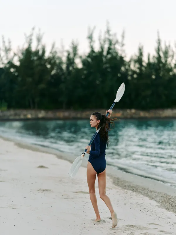 Young swimmer on a sandy beach carries two white paddles, ready for a water sport session. calm sea, soft light, and distant trees frame a peaceful scene.