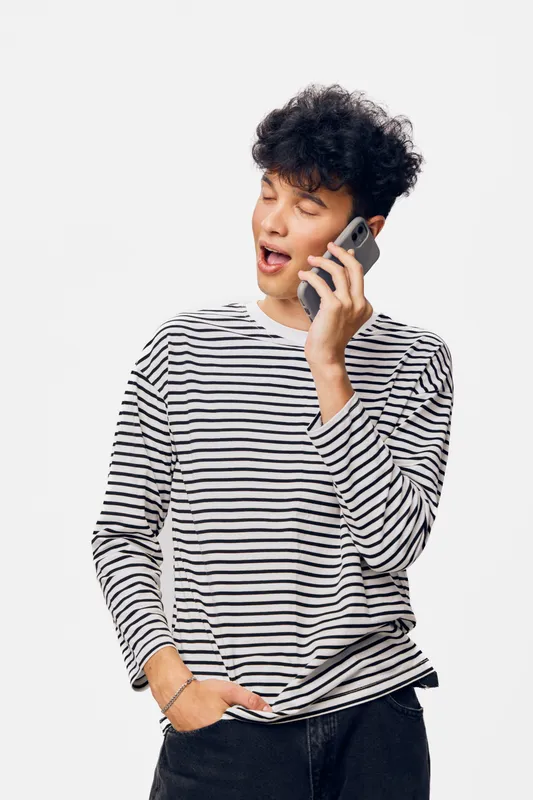 young man talking on smartphone with expressive face wearing striped shirt standing against white background, casual style and communication concept