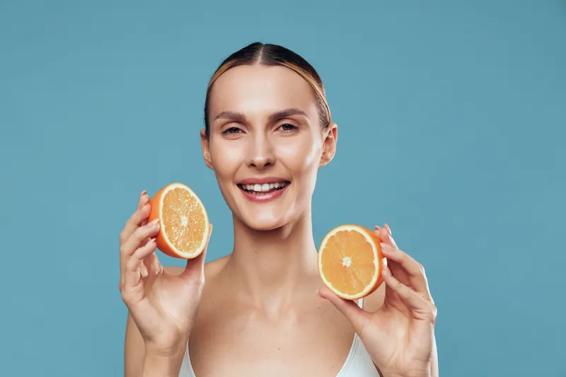 Smiling young woman holds two orange halves against a blue background, fresh citrus for vitamin C, nutrition and healthy lifestyle, wellness and diet concept self care and immunity