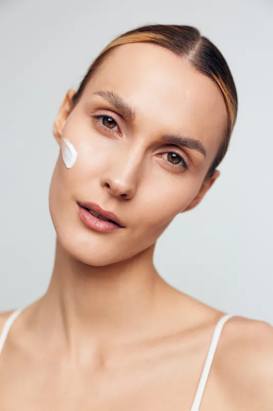 Young woman with clean fresh skin applies white moisturizing cream on cheek looking at camera with calm expression isolated on light background in skincare beauty concept.
