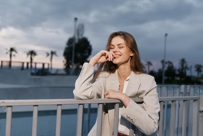 young woman smiling outdoors in beige jacket with relaxed expression during cloudy day in urban setting with palm trees and metal railing