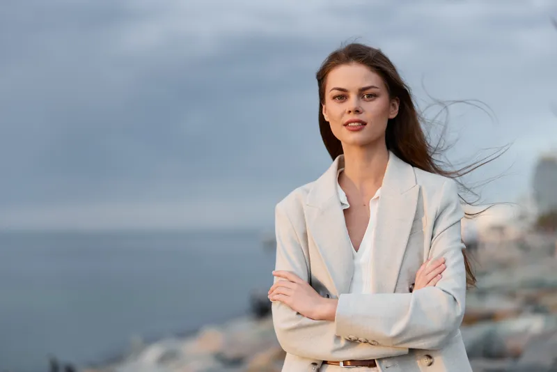 confident woman in light beige blazer standing with arms crossed near sea under cloudy sky with calm expression and natural makeup