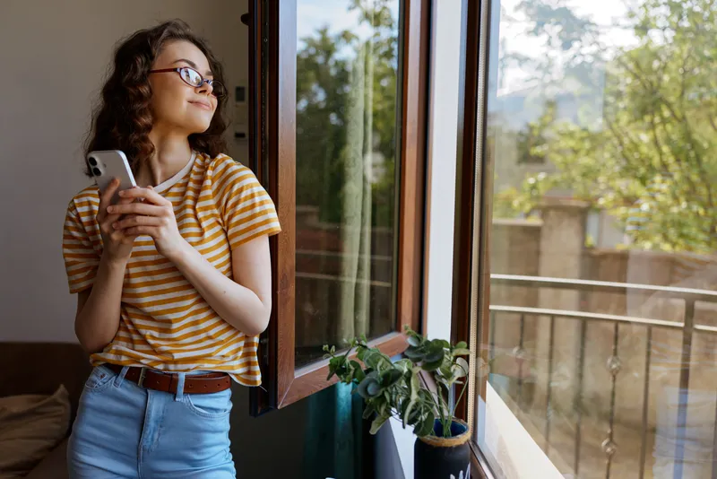 Young woman stands by an open window at home, gazing outside while holding a smartphone, wearing a striped yellow shirt and jeans, with a plant on the sill and bright natural light.