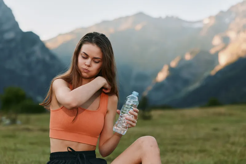 A young woman sits outdoors in a scenic meadow with mountains in the background, holding a water bottle. She wears an athletic top and looks relaxed, refreshed, and carefree.