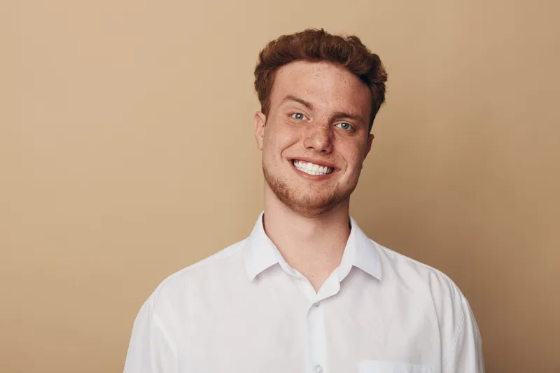 Smiling young man with curly hair wearing white shirt stands against beige background. Portrait of happy confident male model with natural expression and casual style in studio lighting.