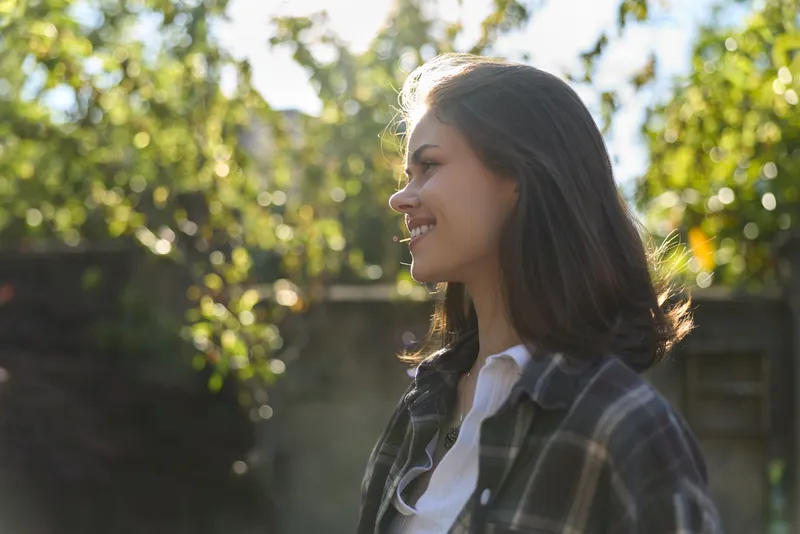 Cheerful woman outdoors, smiling in sunlight, casual plaid shirt, natural lifestyle moment, greenery backdrop, soft breeze, candid portrait, relaxed vibe, warm tones, subtle glow