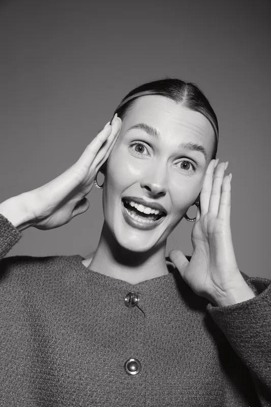 Happy Caucasian woman with fair skin in sweater expresses surprise with hands near face on grey background studio.