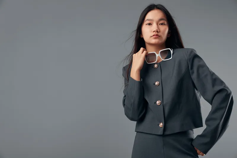 Young woman with long hair wearing dark gray jacket and pants stands against gray background holding white sunglasses near neck. Fashion model with confident pose and minimal makeup.