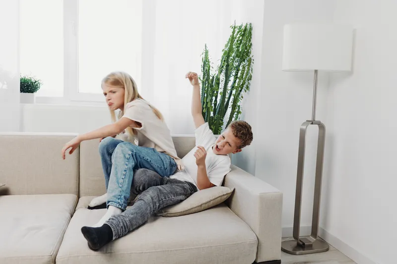 Two children having playful wrestling on a beige sofa in a bright modern living room with natural light. Concept of childhood fun, sibling interaction, and energetic home activities.