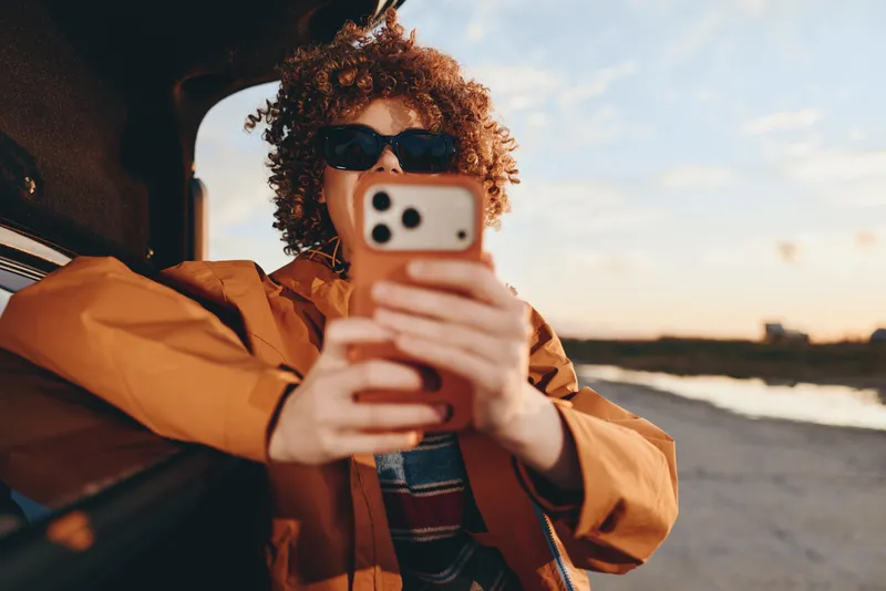 Woman lifestyle at sunset wearing sunglasses and a jacket holding a smartphone near a car on an open road outdoors travel moment with warm sky distant horizon warm evening light getaway