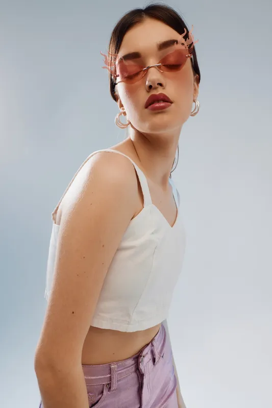 Young woman in a white crop top and pink pants poses confidently wearing pink sunglasses and hoop earrings. Studio shot on a grey background. Concept of woman beauty fashion.