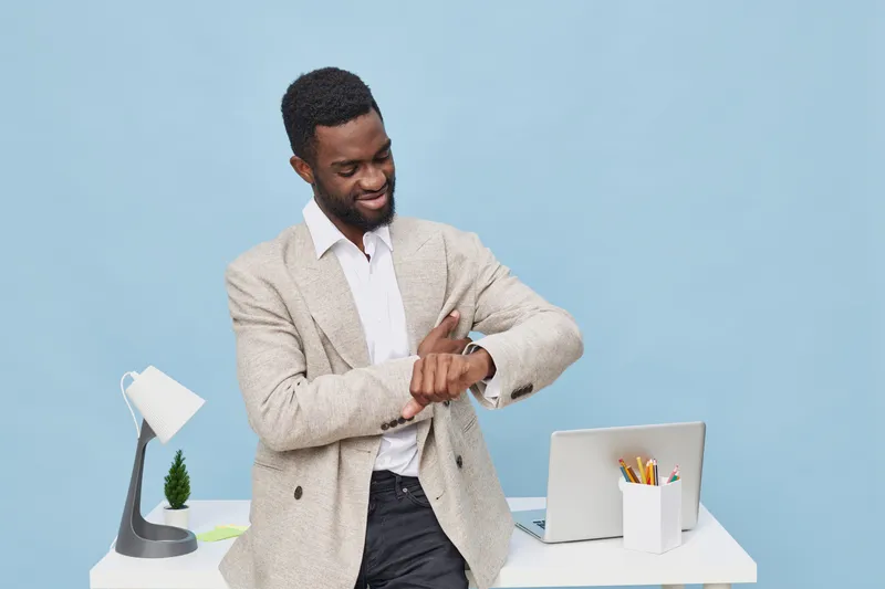 Confident smiling man in smart casual attire checking wristwatch while standing at modern workspace with laptop, desk lamp, and stationery on a blue background. Studio shot. Professional lifestyle con