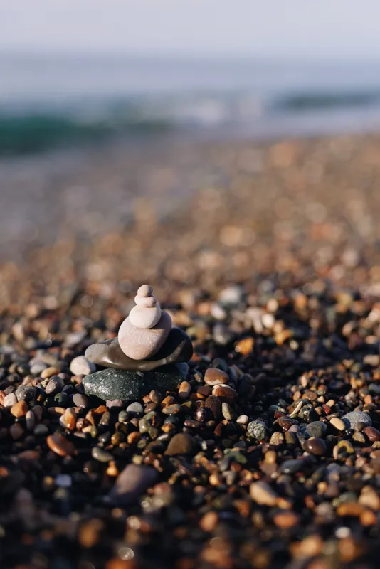 Stacked stones on a pebbled beach with a blurred ocean backdrop, symbolizing balance and tranquility. Close up of a small tower amid varied gravel and seafront breeze.