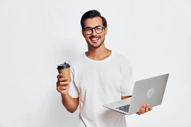 Happy cheerful young man holding a laptop in one hand and a coffee cup in the other, smiling at the camera against a plain white background. Work lifestyle concept.