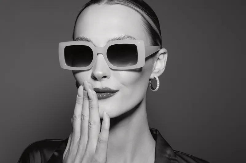 Stylish young caucasian woman with fair skin wearing large sunglasses and hoop earrings poses with hand near mouth. Black and white portrait with elegant and modern fashion concept.