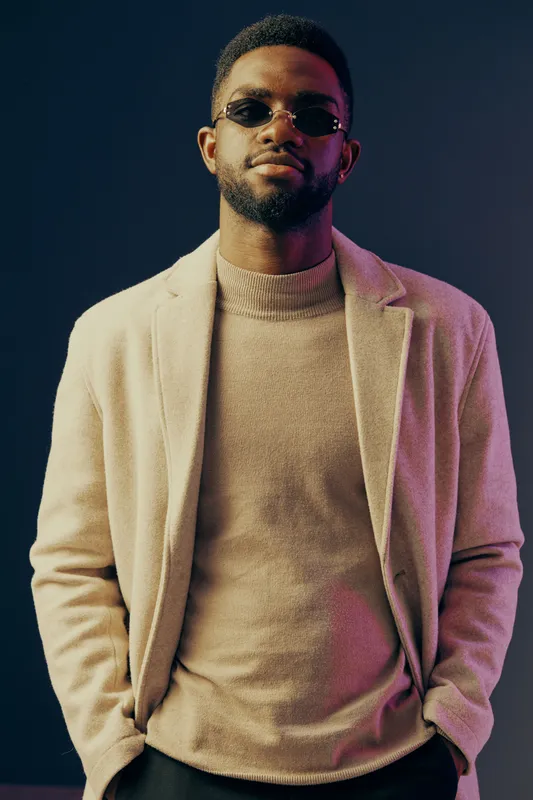 Stylish African man in sunglasses and beige coat, posing confidently against a colorful backdrop, showcasing modern fashion and urban lifestyle