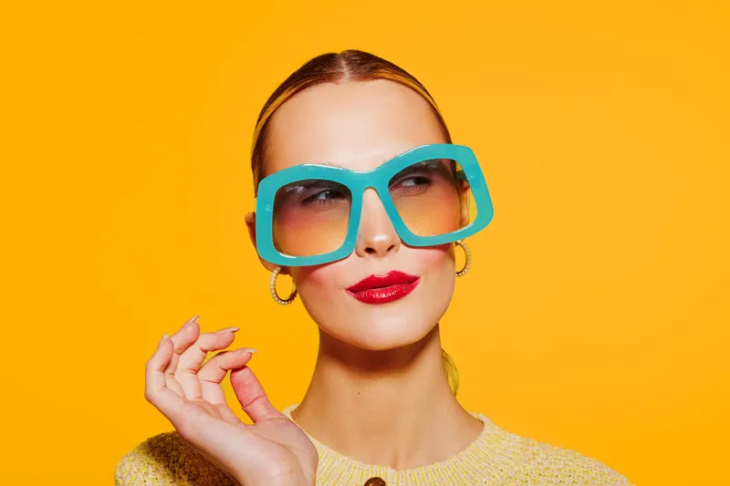 Stylish woman with large blue sunglasses and red lipstick poses against bright orange background. Fashion portrait with confident expression and trendy accessories.