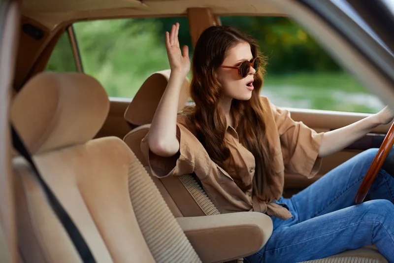 Young woman in casual outfit expressing joy while driving in a vintage car, surrounded by nature, showcasing vibrant colors and a carefree summer vibe.