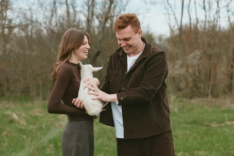 A smiling couple in casual outdoor clothing cradle a baby goat kid in a sunlit field, capturing a warm moment of family life, affection, and gentle animal companionship.