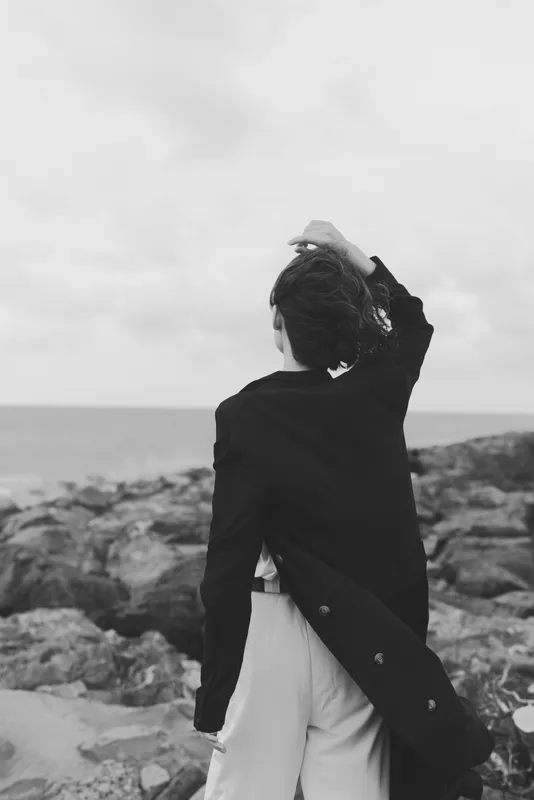 Woman coastline rocks jacket backview ocean windblown seaside fashion solitude, a monochrome mood captures a reflective moment along rugged shore with the sea at the horizon