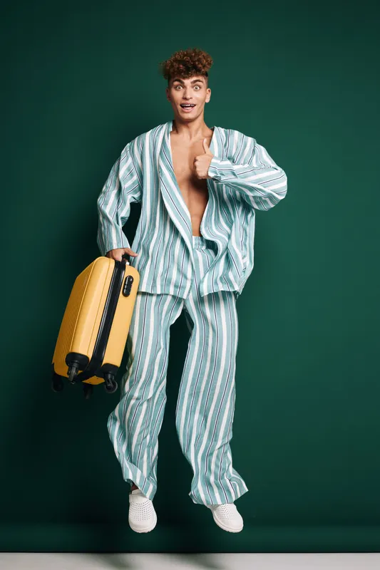 Young man in striped pajamas holding yellow suitcase jumping with excitement on green background. Casual travel, leisure and happy expression for vacation or trip preparation.