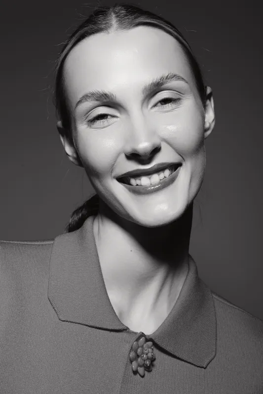 Portrait of a smiling Caucasian woman with fair skin and pulled-back hair wearing a textured top with a decorative brooch on a dark background.