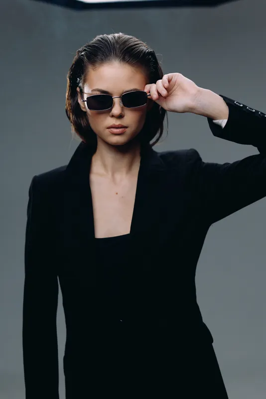 Stylish woman wearing black sunglasses and blazer with confident expression on gray background