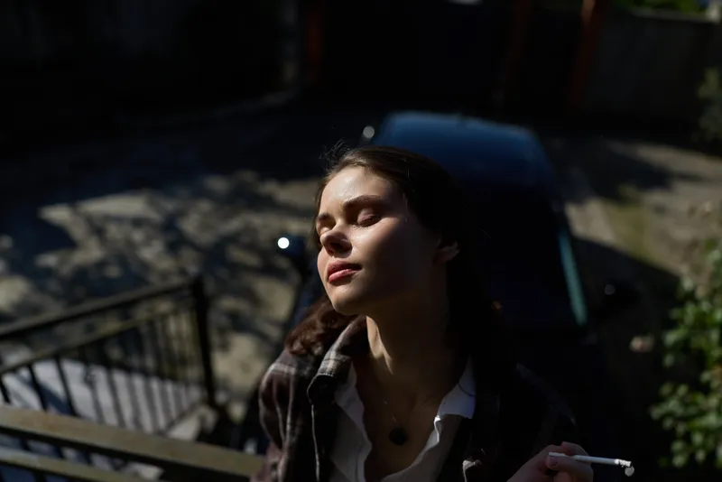 Calm woman outdoors, sunlit portrait with eyes closed, relaxed posture on a balcony, soft breeze, natural colors, urban background, casual style, lifestyle moment, graceful composition