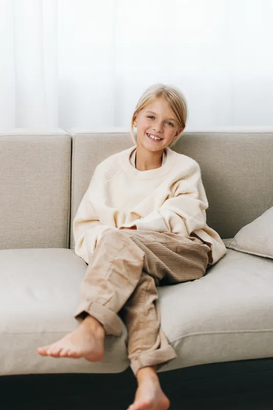 Authentic lifestyle image of a child at home, relaxed on a sofa in warm casual wear, playful mood, school mindset, everyday comfort and a natural smile.