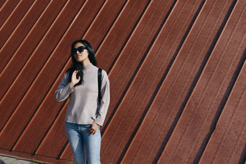 Woman wearing sunglasses and backpack stands against a diagonal wooden wall in casual street fashion, outdoors, urban setting, relaxed pose, daytime light.