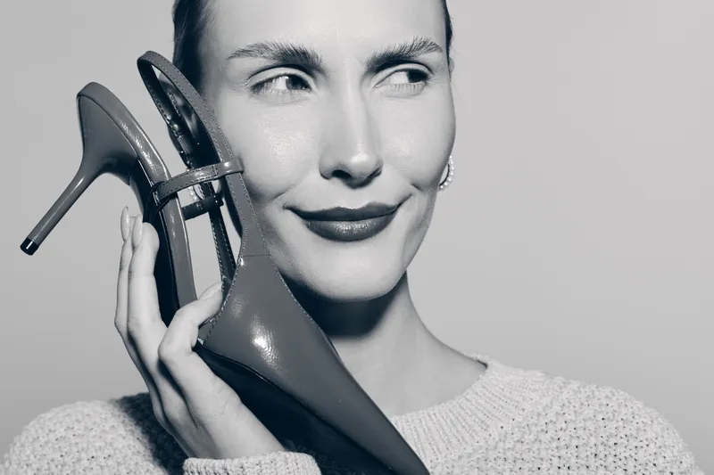 Portrait of a fair skinned caucasian woman holding high heel shoe near face, smiling subtly. Black and white studio image, fashion and beauty concept.