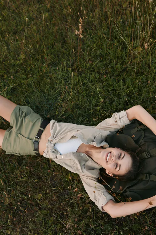 Young woman lies on grass outdoors, smiling with arms raised, wearing a light jacket and shorts, enjoying a relaxed moment in warm afternoon light