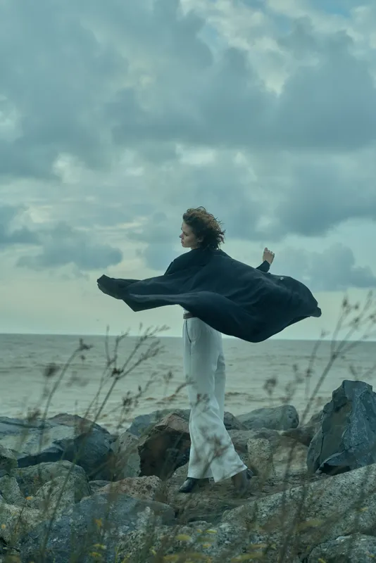 Woman stands on a rocky shore by the sea, cape flowing in the wind, capturing movement, freedom and a dramatic landscape in outdoor nature mood