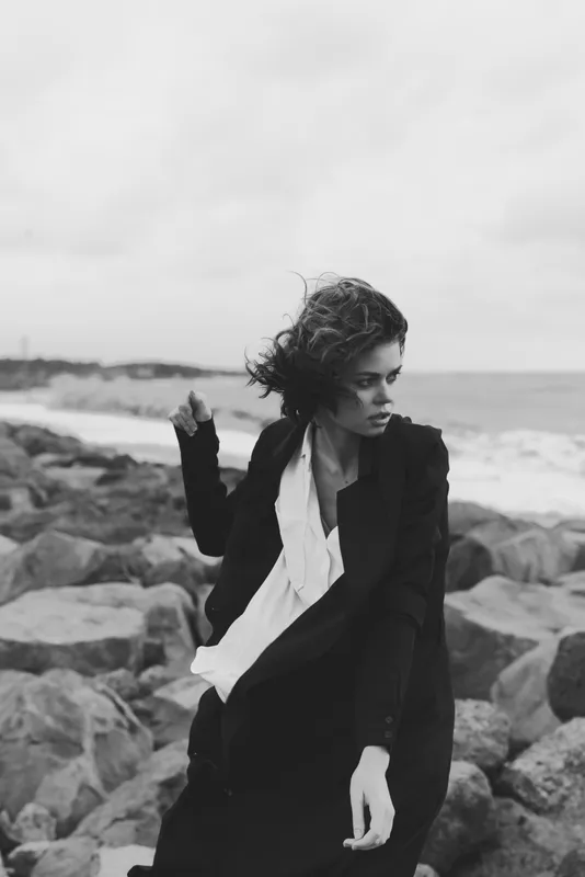 Fashionable woman on a windswept shore among rocks, wearing a blazer and scarf in black and white, delivering editorial style, dramatic mood and seaside elegance for photography