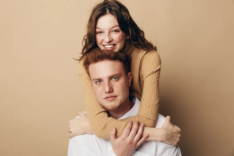 Smiling woman with curly hair hugs serious man from behind on beige background. Young couple shows affection and connection in casual clothing. Concept of love, relationship, and friendship.