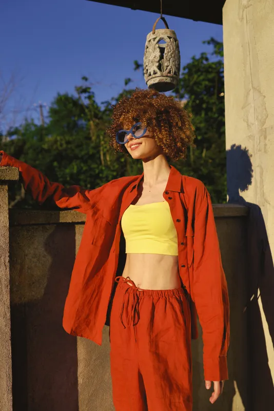 A stylish woman with curly hair in a bright red two piece set stands outdoors, wearing sunglasses, posing confidently in sunlit urban surroundings
