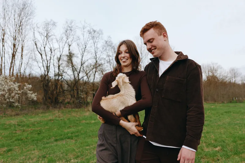Cheerful couple outdoors, smiling at each other, small dog cradled by the woman, casual outfits, sunlit park scene, relaxed mood, natural colors, lush grass, gentle breeze, candid moment, warm