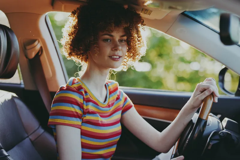 Cheerful woman driving a car, wearing a colorful striped shirt, sunlight through the window, candid lifestyle moment, relaxed pose, warm retro color palette