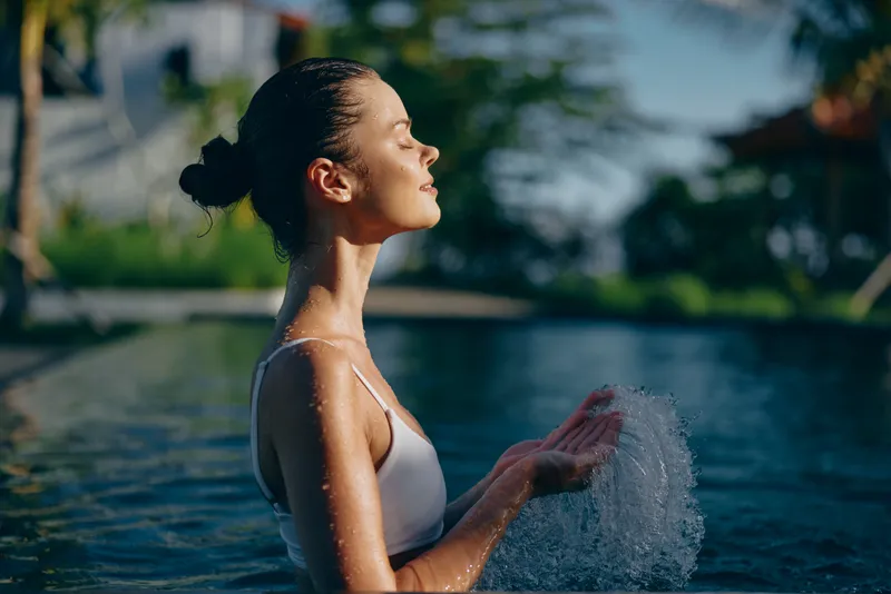Young woman in white swimsuit standing in pool with closed eyes, enjoying sunlight and splashing water, relaxed mood, natural outdoor setting with greenery background