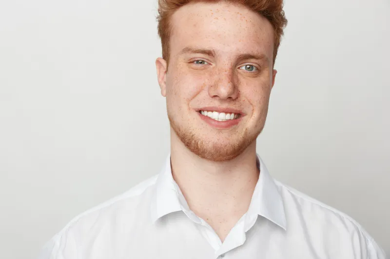 Smiling young man with red hair and beard wearing white shirt looks at camera. Studio portrait on light background. Concept of confidence, happiness, and casual style for men.
